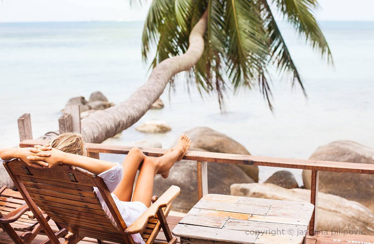 Marina from Health Glows resting on a balcony with a view of a palm tree and the sea on the Thai island Koh Phangan. The picture is part of the 'Health Glows Thai Retreat' promo page.