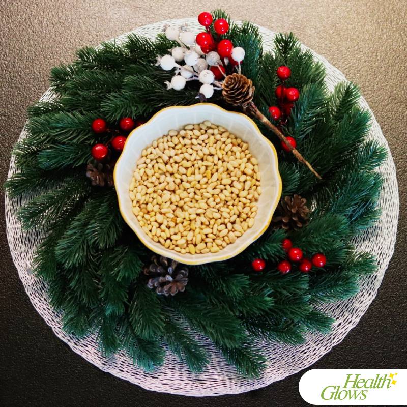 Raw pine nuts in a bowl that is surrounded with a decorative evergreen branch with white and red and white berries.