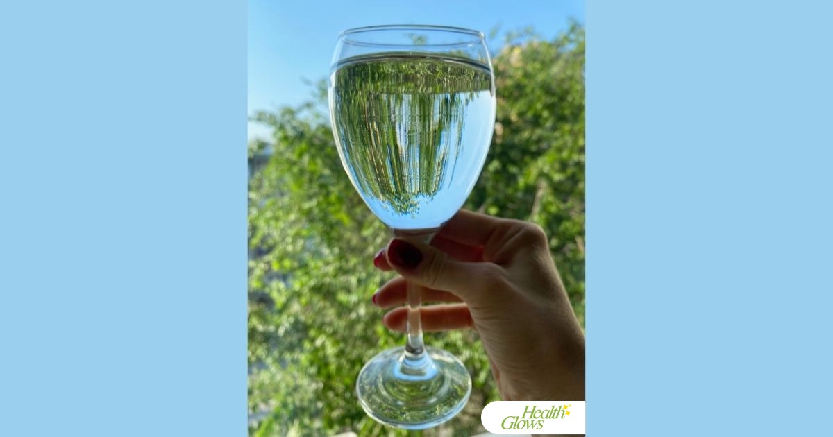 A woman's hand holding a wine glass full of clear water in front of a window. There are a green treetop and blue sky behind the glass.