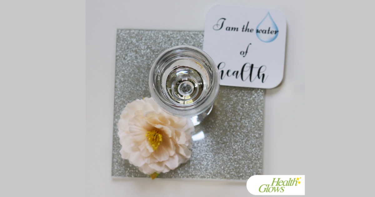 A wine glass filled with pure water on a silver coaster, with a pale pink flower and a coaster that says 'I Am the Water of Health' - pictured from above.
