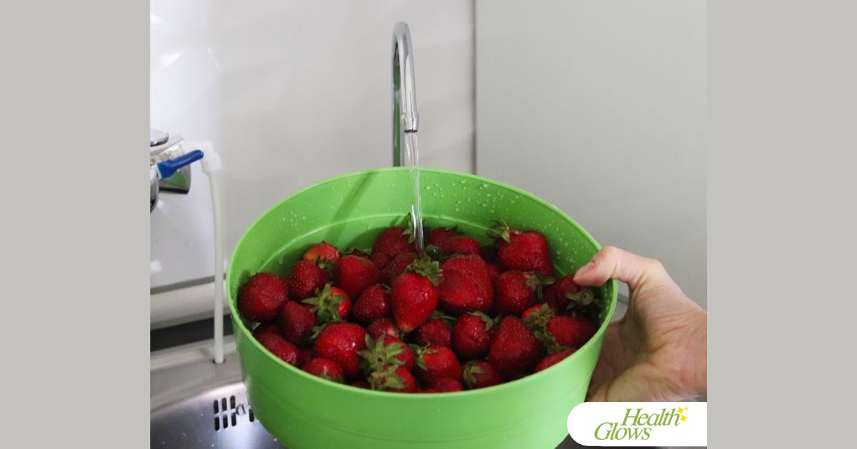 A woman's hand holding a green sieve full of strawberries under a stream of water running from a water filter tap.