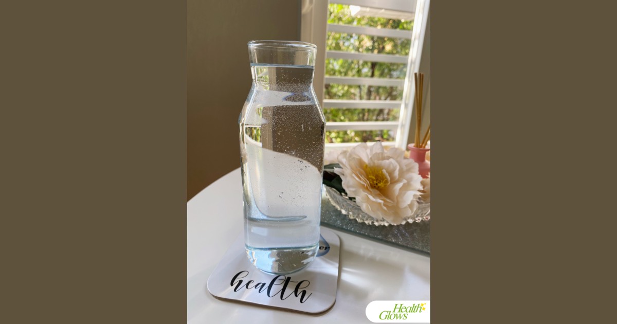 A glass bottle with clean water standing on a coaster that reads 'I Am the Water of Health'. Next to it is a silver coaster with a decorative flower in a glass bowl.