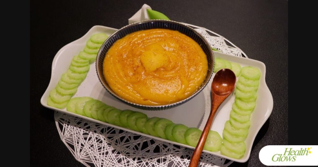 An example of a raw vegan lunch. Persimmon pudding and cucumber slices, with a wooden spoon, a napkin in a decorative tulip holder, on a white tray, a silver table mat, and a black table.