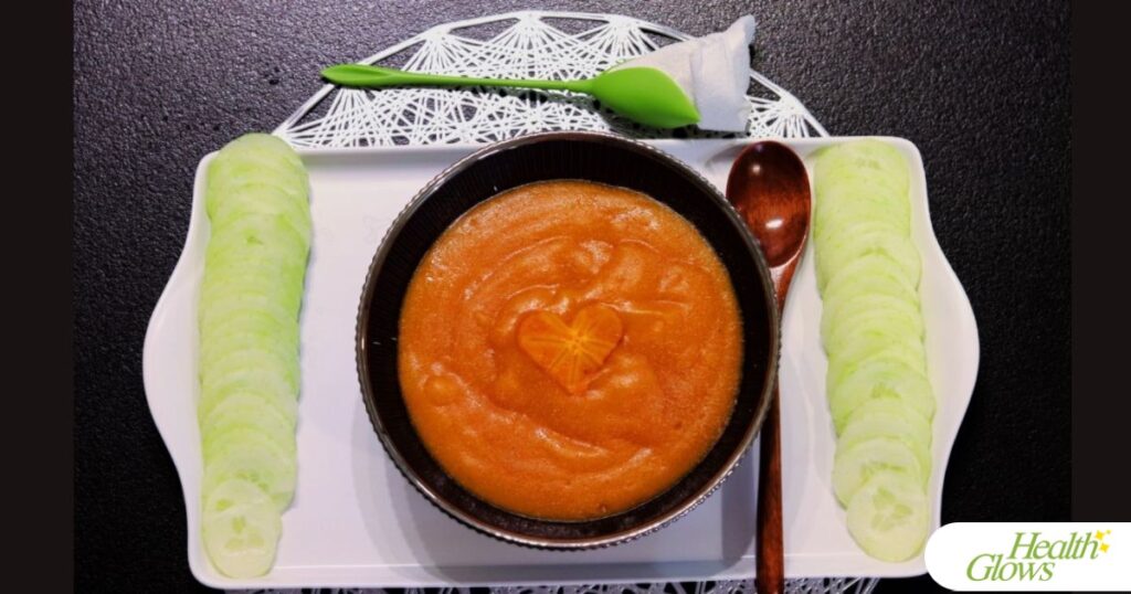 Persimmon pudding in a blue bowl, a wooden spoon, cucumber slices, a napkin in decorative tulip holder, a white tray, a black table.