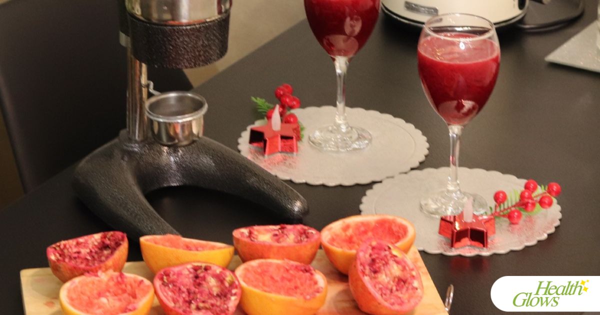 Halved and juiced pomegranates, oranges and grapefruits laid out next to a pomegranate press. On the other side of the pomegranate press there are two wine glasses filled with pomegranate juice.