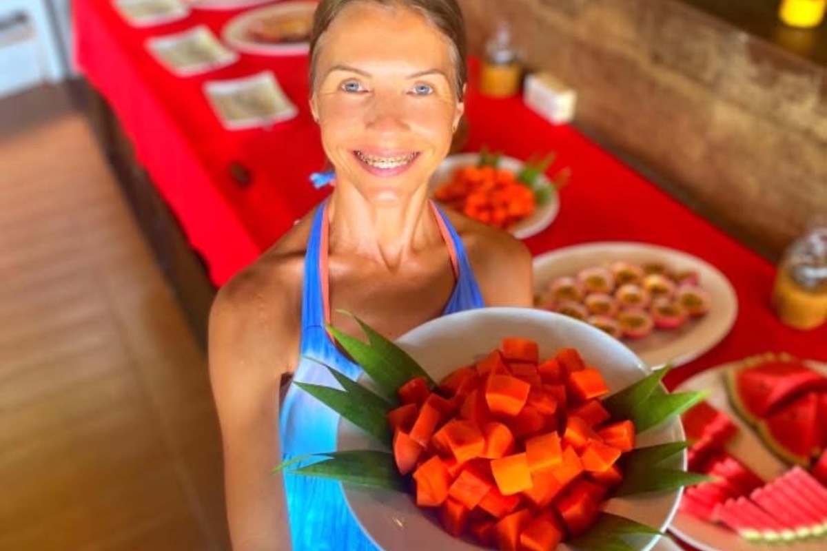 Marina from Health Glows standing next to a raw vegan breakfast buffet, holding a plate filled with diced papaya and smiling into the camera.
