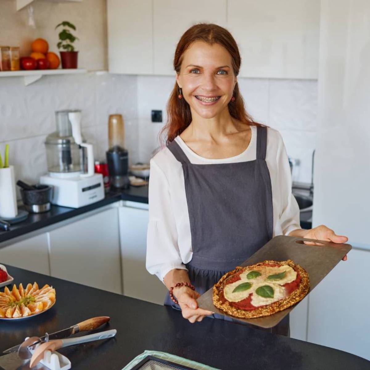 Marina from Health Glows is holding a dehydrator shelf with a raw vegan pizza on it, standing in her stove-free kitchen.